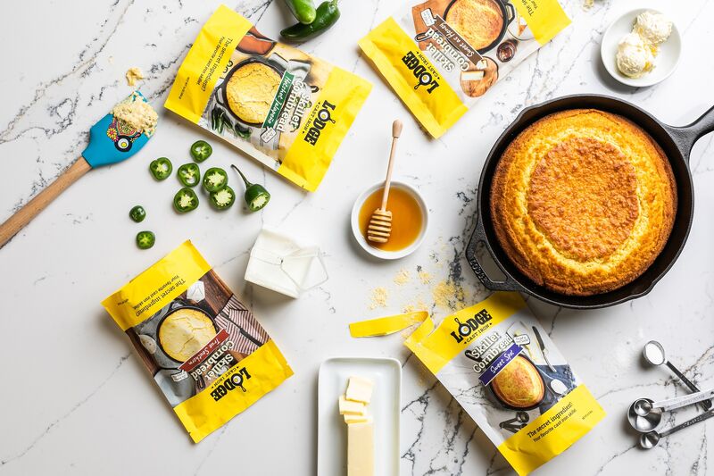 An image showcasing Lodge's brand licensing skillet cornbread mixes, featuring an assortment of their branded products thoughtfully arranged on a tabletop. Each cornbread mix package prominently displays the Lodge logo, ensuring the unmistakable quality and authenticity associated with the brand. Next to the mixes, a well-seasoned Lodge cast iron skillet sits alongside, reflecting the ideal pairing for creating mouthwatering cornbread. Fresh corn kernels add a touch of farm-fresh appeal to the scene, reinforcing the wholesome, home-cooked goodness that Lodge brings to every meal. OWS Foods used Broad Street Licensing Group to achieve thier goal in licensing the Lodge brand.