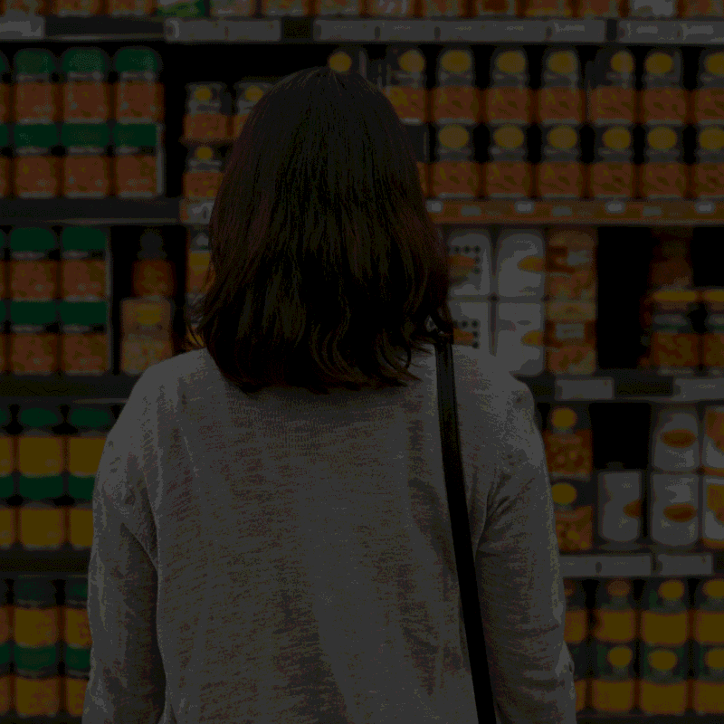 Woman browsing canned food products in a grocery store aisle, highlighting consumer choice and familiar food brands.