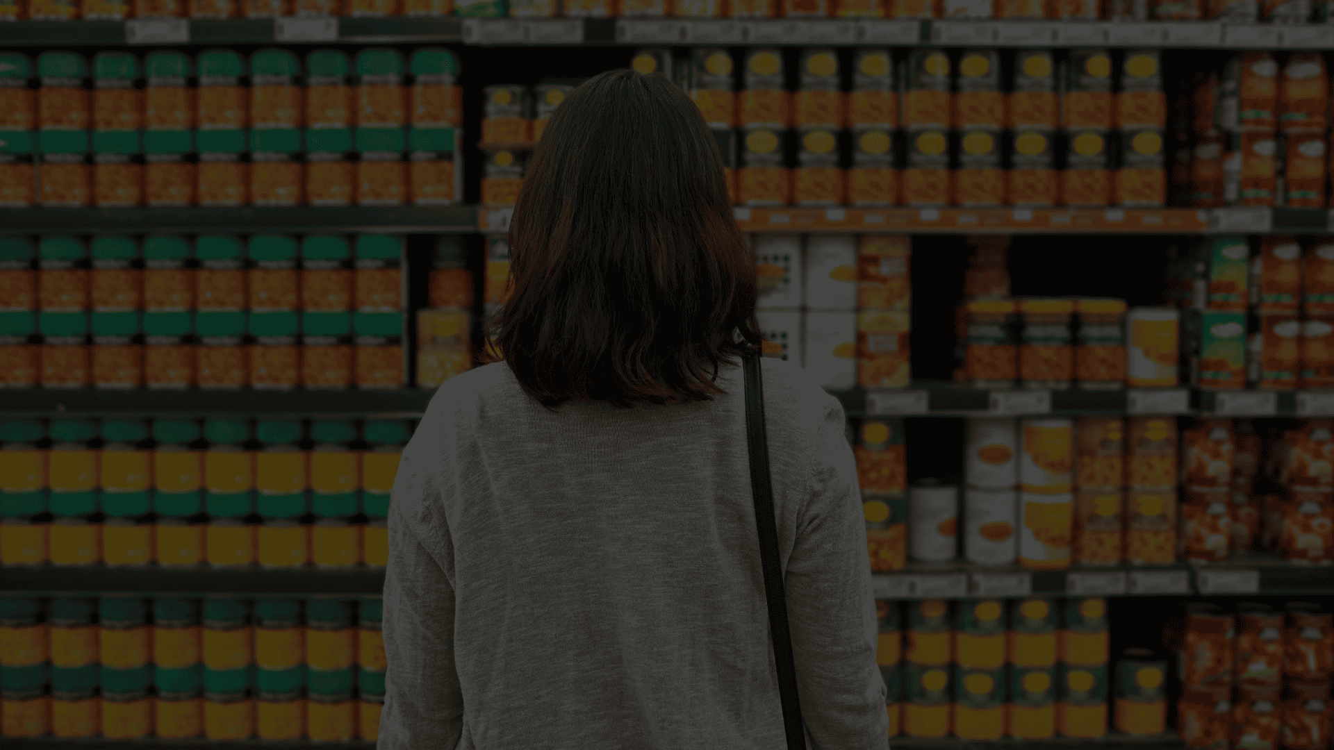 Woman browsing canned food products in a grocery store aisle, highlighting consumer choice and familiar food brands.